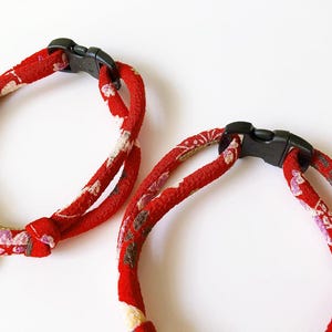 May include: Two red fabric pet collars with black plastic buckles. The collars have a floral pattern in white, pink, and gold. One collar has a gold-colored charm with a carved design.
