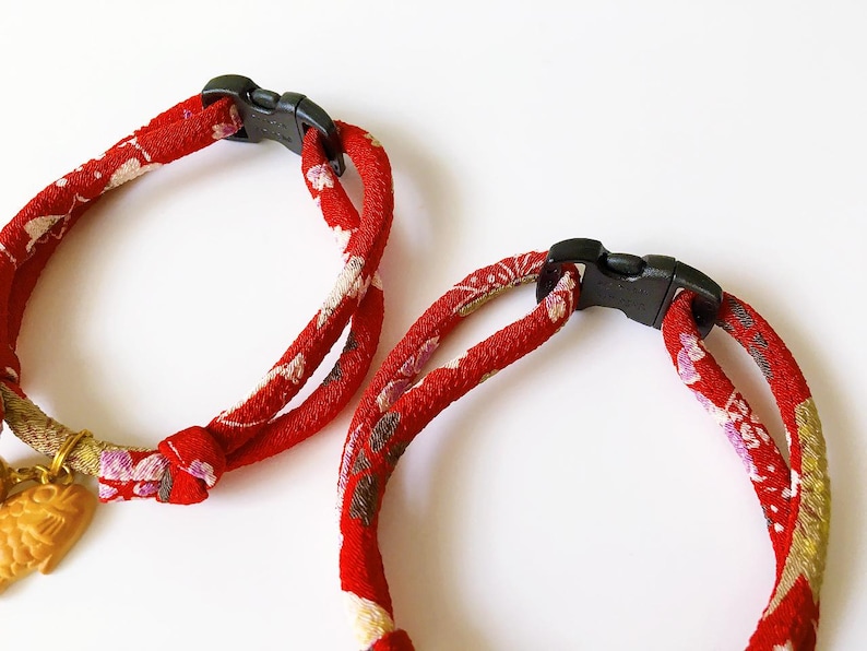 May include: Two red fabric pet collars with black plastic buckles. The collars have a floral pattern in white, pink, and gold. One collar has a gold-colored charm with a carved design.