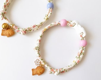 May include: Two floral patterned cat collars. One collar has a gold bell and a fish charm. The other collar has pink beads and a white buckle. The collars are white with a floral pattern of pink and yellow flowers.