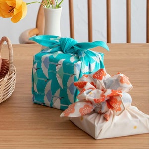 May include: Two gifts wrapped in fabric. One gift is wrapped in a turquoise fabric with a white crane pattern. The other gift is wrapped in a white fabric with a red and orange floral pattern.