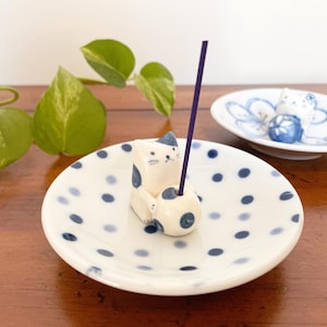 May include: A white ceramic incense holder featuring a cat design with blue accents, holding a purple incense stick. The holder sits on a white plate with blue polka dots. Another small dish with a cat and floral design is in the background.