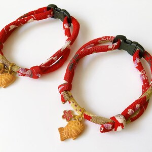 May include: Two red cat collars with a floral pattern and black plastic buckles. Each collar has a gold-colored fish charm and a small bell. The collars are designed for pets.