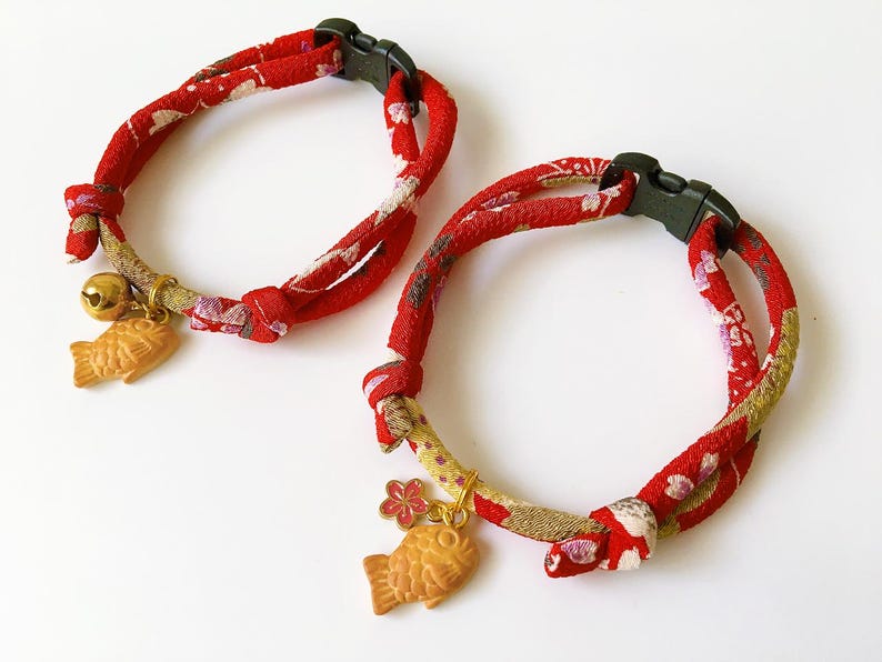 May include: Two red cat collars with a floral pattern and black plastic buckles. Each collar has a gold-colored fish charm and a small bell. The collars are designed for pets.