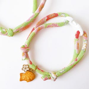 May include: Two cat collars with a floral pattern in green, pink, and orange. Each collar has a gold-colored fish charm and a white plastic clasp. The collars are knotted and appear to be made of fabric.