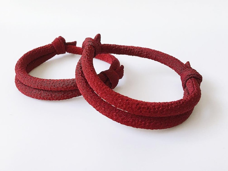 May include: Two red fabric bracelets with a knotted design. The bracelets are a deep burgundy color and appear to be made of a woven material. The bracelets are displayed on a white background.