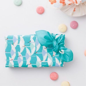 May include: A turquoise gift box with a white crane pattern and a turquoise ribbon tied in a bow. The box is surrounded by pastel-colored candy.