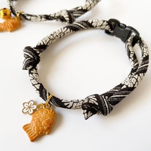 May include: Two cat collars with a black and white patterned design. Each collar has a gold-colored charm, one shaped like a fish and the other a flower. One collar has a small gold bell.
