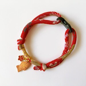 May include: Red and gold patterned fabric pet collar with a black plastic buckle. The collar features a small gold charm with a fish and flower design. The collar is designed for a small pet.