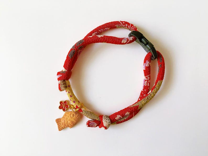May include: Red and gold patterned fabric pet collar with a black plastic buckle. The collar features a small gold charm with a fish and flower design. The collar is designed for a small pet.