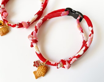 May include: Two red and white patterned fabric collars with gold charms. One collar has a bell and a gold fish charm. The other collar has a gold flower and a gold fish charm.