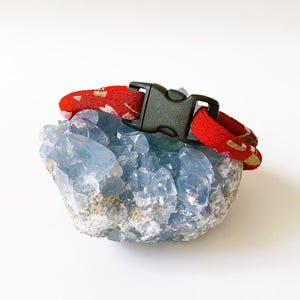 May include: A red patterned collar with a black buckle rests on a light blue crystal formation. The collar has a repeating gold and white design. The crystal has a rough, textured surface and is a pale blue color.