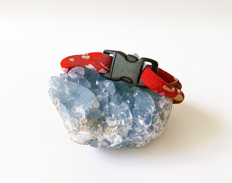 May include: A red patterned collar with a black buckle rests on a light blue crystal formation. The collar has a repeating gold and white design. The crystal has a rough, textured surface and is a pale blue color.
