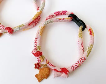 May include: Two pink and floral patterned pet collars with a black clasp. Each collar has a gold-colored fish charm and a red flower charm. The collars are made of a soft, patterned fabric with red, yellow, and green accents.