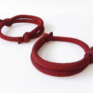 May include: Two red bracelets with a knotted design. The bracelets are made of a textured material and are circular in shape. The knots are located on the top of the bracelets. The background is white.