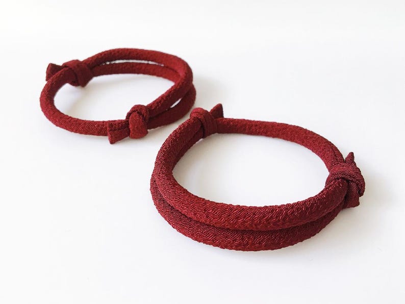 May include: Two red bracelets with a knotted design. The bracelets are made of a textured material and are circular in shape. The knots are located on the top of the bracelets. The background is white.