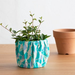 May include: A small green plant in a turquoise fabric pot with a white crane pattern. The pot is sitting on a light brown wooden surface. A terracotta pot is sitting to the right of the fabric pot.