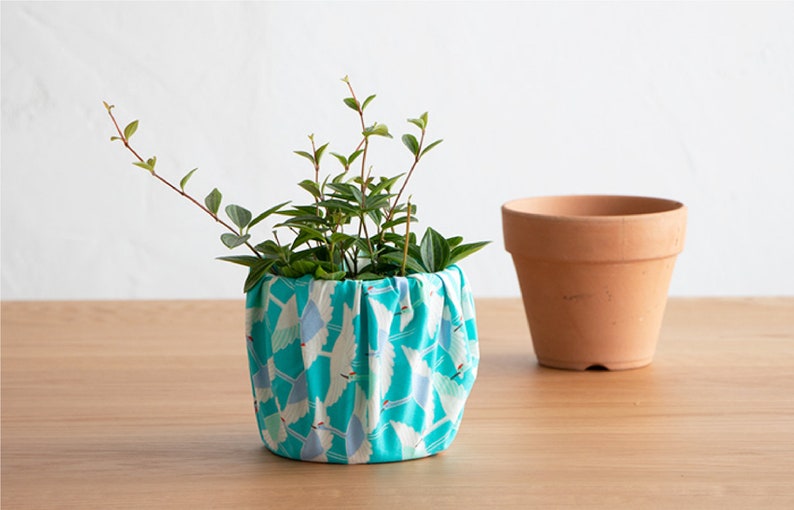 May include: A small green plant in a turquoise fabric pot with a white crane pattern. The pot is sitting on a light brown wooden surface. A terracotta pot is sitting to the right of the fabric pot.