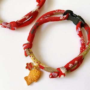May include: Two red and patterned cat collars with black plastic buckles. Each collar features a gold-colored charm, one with a fish design and the other with a bell. The collars are made of a patterned fabric with red, white, and gold hues.