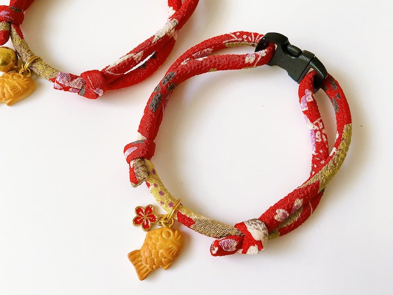 May include: Two red and patterned cat collars with black plastic buckles. Each collar features a gold-colored charm, one with a fish design and the other with a bell. The collars are made of a patterned fabric with red, white, and gold hues.