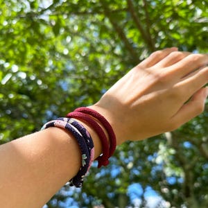 May include: Three bracelets on a wrist. One is navy blue with a floral pattern, and two are solid burgundy. The bracelets are made of a rope-like material. The background is blurred green foliage and blue sky.