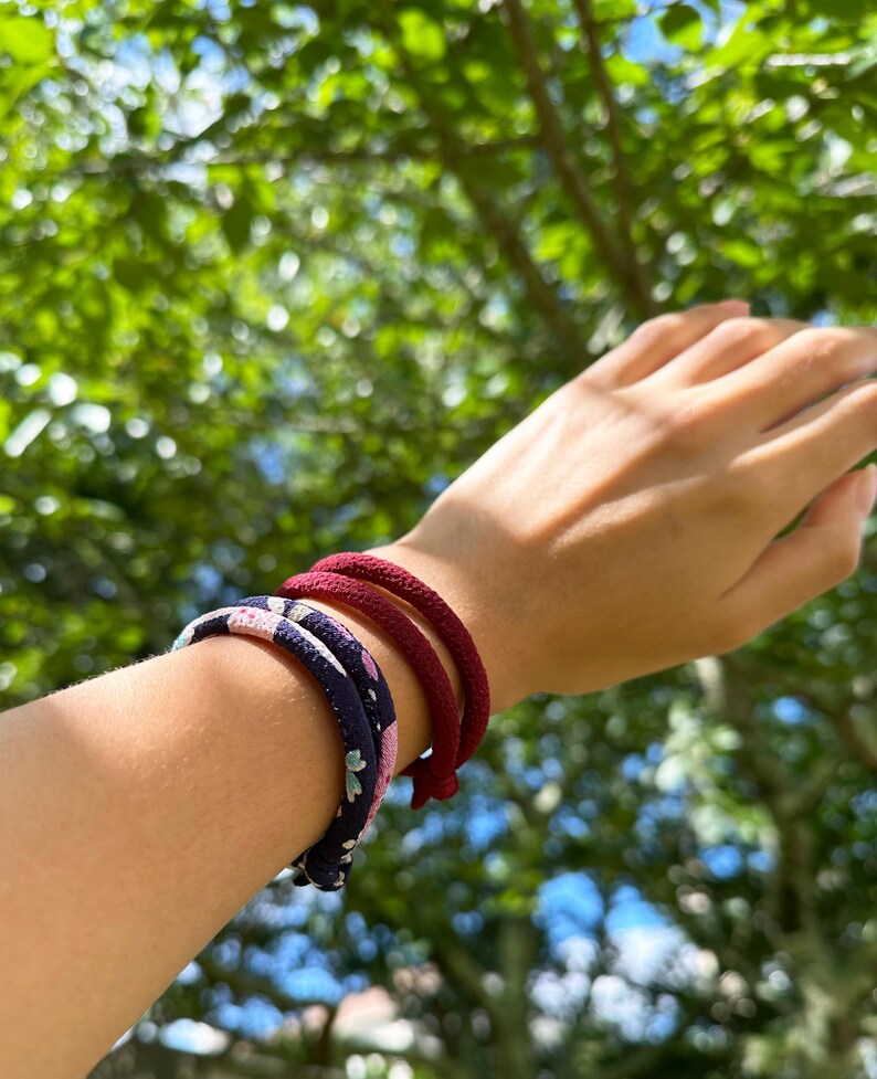 May include: Three bracelets on a wrist. One is navy blue with a floral pattern, and two are solid burgundy. The bracelets are made of a rope-like material. The background is blurred green foliage and blue sky.