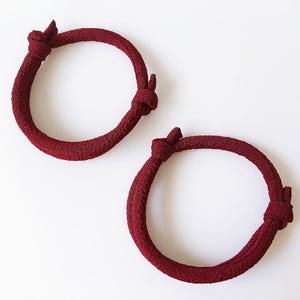 May include: Two burgundy-colored rope bracelets with a knotted design. The bracelets are circular and rest on a white surface. The rope has a textured appearance, and each bracelet has two knots.