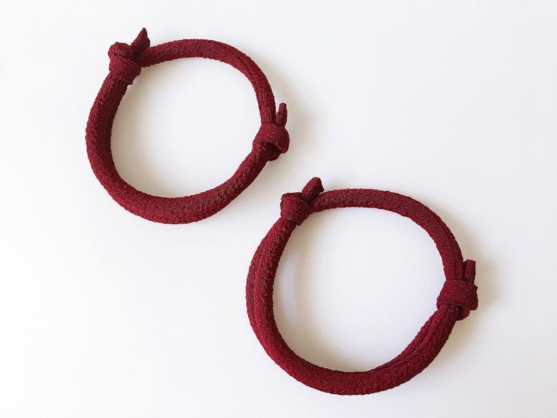 May include: Two burgundy-colored rope bracelets with a knotted design. The bracelets are circular and rest on a white surface. The rope has a textured appearance, and each bracelet has two knots.