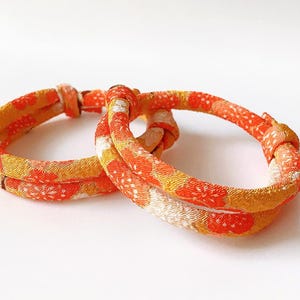 May include: Two orange and yellow fabric bracelets with a floral pattern. The bracelets are knotted and appear to be made of a soft, textured material. The background is plain white.