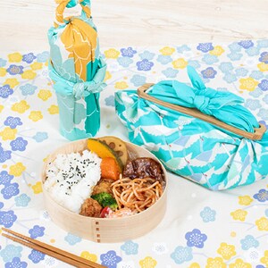 May include: A wooden lunchbox with a blue and white floral design, filled with rice, vegetables, and meat. The lunchbox is wrapped in a blue and white fabric with a crane pattern. Two chopsticks are on the table next to the lunchbox.