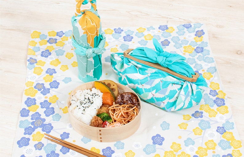 May include: A wooden lunchbox with a blue and white floral design, filled with rice, vegetables, and meat. The lunchbox is wrapped in a blue and white fabric with a crane pattern. Two chopsticks are on the table next to the lunchbox.