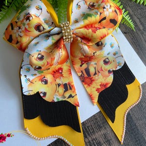 May include: A large, decorative hair bow featuring a bee-themed design. The bow has a yellow base with a black accent and is adorned with a repeating pattern of cartoon bees and flowers. A rhinestone clasp secures the bow's center.