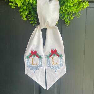 Peut inclure: Décoration de porte blanche avec des détails brodés. Comprend un nœud rouge, une couronne verte et un motif de treillis bleu clair avec la lettre "L" en or. Le porte-manteau est suspendu à une couronne verte.