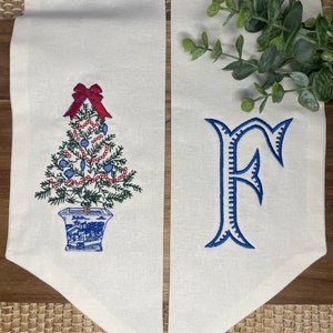 Peut inclure: Deux fanions en tissu blanc avec des broderies bleues. Le fanion de gauche représente un sapin de Noël bleu et blanc avec des ornements rouges et un nœud rouge. Le fanion de droite représente une lettre "F" brodée en bleu.