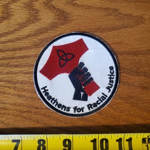 May include: White embroidered patch with a red hammer and a black fist holding it. The hammer has a black triquetra symbol on it. The text around the image reads "Heathens for Racial Justice".