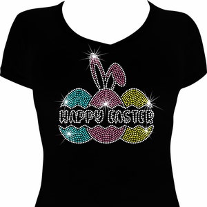 May include: A black short-sleeved t-shirt featuring a rhinestone graphic of three Easter eggs in turquoise, pink, and yellow. The design includes white rhinestone bunny ears on top and the text HAPPY EASTER in white rhinestones across the center.