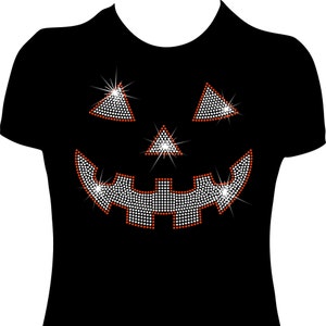 Puede incluir: Camiseta negra con un diseño de cara de calabaza de strass. La calabaza tiene una amplia sonrisa y ojos naranjas.