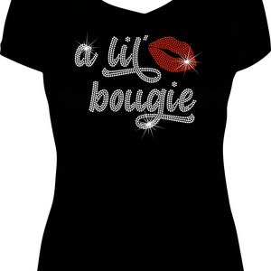 May include: A black short-sleeved t-shirt with a V-neckline featuring a rhinestone graphic on the chest. The design consists of the text 'a lil' bougie' in white rhinestones and a red rhinestone lip icon, creating a sparkling, textured effect.