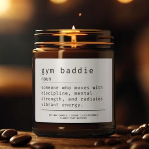 Puede incluir: Una vela de cera de soja encendida en un frasco de vidrio marrón con las palabras "gym baddie" en una etiqueta blanca. La etiqueta define "gym baddie" como alguien que se mueve con disciplina, fuerza mental y energía vibrante. Granos de café esparcidos.