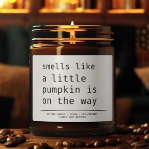 Puede incluir: Una vela en tarro de cristal marrón con una mecha encendida. La etiqueta dice "smells like a little pumpkin is on the way". La vela está rodeada de granos de café. Cera de soja, vegana y ecológica.