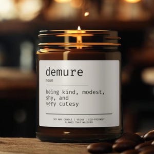 Puede incluir: Un tarro de vela de vidrio marrón con una etiqueta blanca que dice "demure noun being kind, modest, shy, and very cutesy SOY WAX CANDLE | VEGAN | ECO-FRIENDLY FLAMES THAT WHISPER".