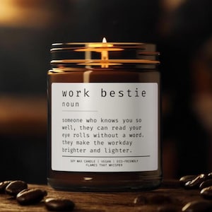 Puede incluir: Vela de tarro de vidrio marrón con tapa dorada, etiquetada como "work bestie". La etiqueta define el término. La vela está encendida y los granos de café están esparcidos alrededor de la base. Hecha de cera de soja.