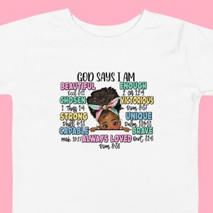 Puede incluir: Una camiseta blanca con un diseño gráfico colorido que presenta a una niña con afro y el texto "Dios dice que soy" con una lista de afirmaciones positivas y versículos bíblicos.