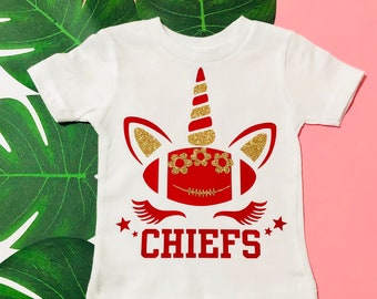 May include: White t-shirt with a red and gold unicorn design. The unicorn's horn is gold with red stripes. The unicorn's eyes are gold glitter with red flowers. The unicorn's face is a red football with white laces. The text "CHIEFS" is in red letters below the unicorn.
