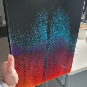 May include: Abstract painting on canvas with a black background and a gradient of colours from blue to purple to red and yellow. The paint is applied in a splatter technique, creating a textured effect.