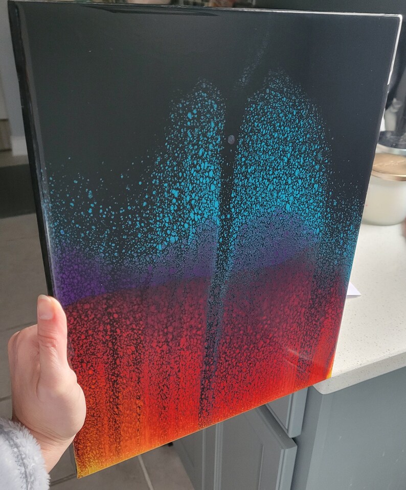 May include: Abstract painting on canvas with a black background and a gradient of colours from blue to purple to red and yellow. The paint is applied in a splatter technique, creating a textured effect.