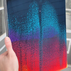 May include: Abstract art painting on a canvas with a black background and a gradient of blue, purple, red, orange, and yellow colours. The paint has a speckled texture.