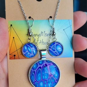 May include: A silver necklace and earrings set with blue and purple abstract art resin pendants. The pendant is round and the earrings are small and round.