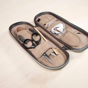 Könnte beinhalten: Geöffnete, mit Reißverschluss versehene Tasche mit verschiedenen Werkzeugen zum Fliegenfischen. Die Tasche ist beige mit einem dunkelbraunen Reißverschluss und Besatz. Im Inneren befinden sich eine Schere, ein Holzwerkzeug und weiteres Angelzubehör. Die Tasche ist geöffnet und zeigt den Inhalt.