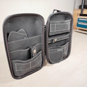 May include: Open, dark grey zippered organiser with multiple pockets and compartments. White stitching accents the pockets and edges. The organiser has a handle and a label that reads "S.Etoja".