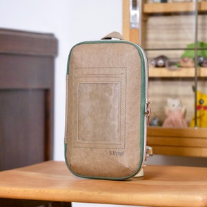 Könnte beinhalten: Eine braune Ledertasche mit einem grünen Reißverschluss und einem grünen, gesteppten Rand. Die Tasche hat ein kleines rechteckiges Fenster auf der Vorderseite. Die Tasche ist mit "5.Etage" beschriftet.